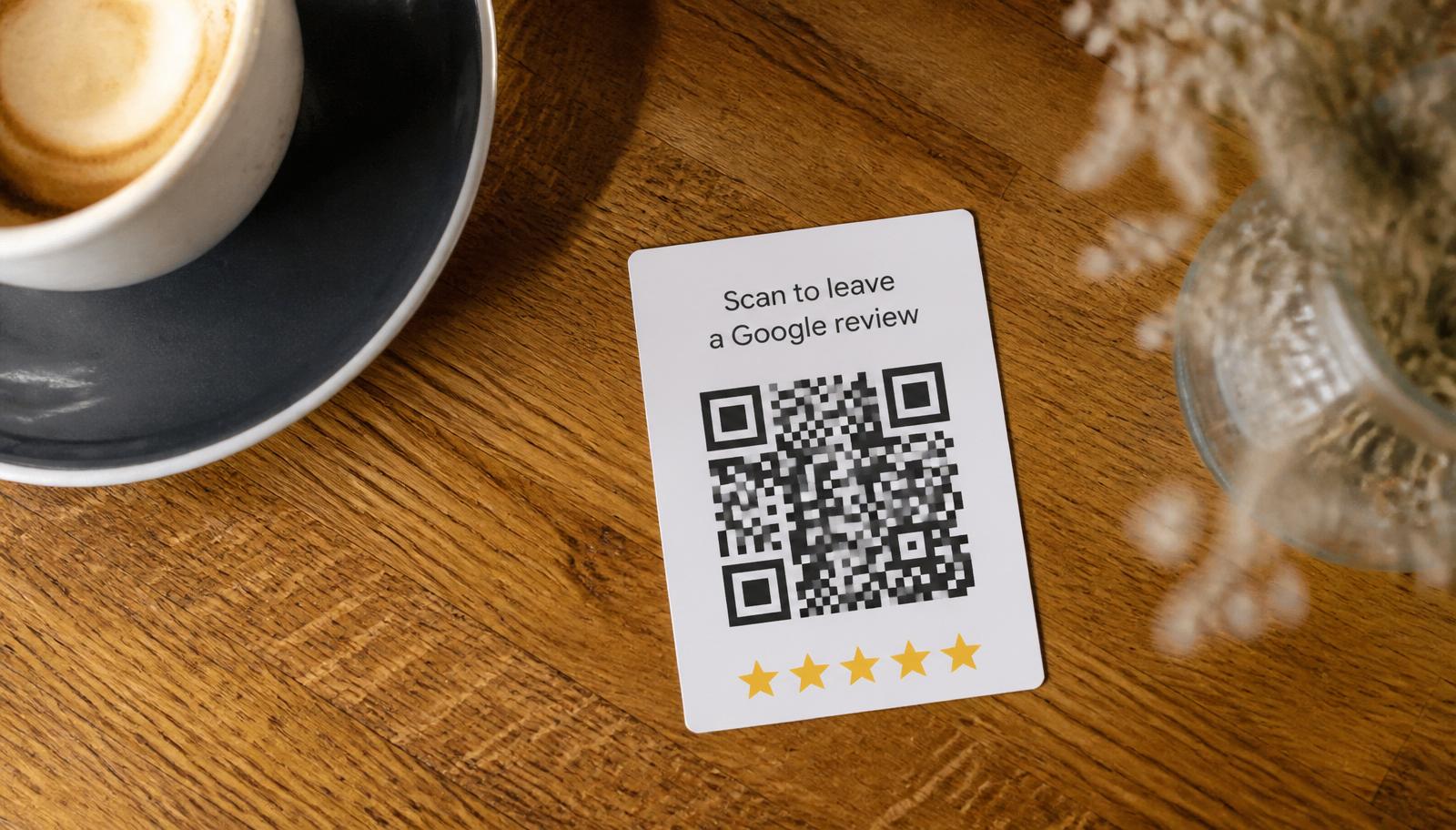 Small printed table talker on a wooden café table next to a coffee cup, showing a black QR code with the copy Scan to leave a Google review and a row of five gold stars